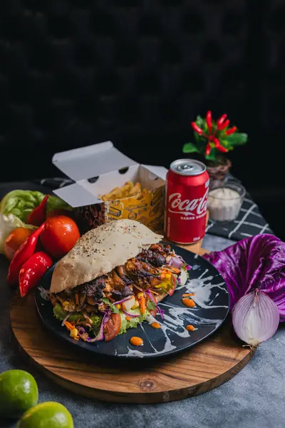 Sanduíche de kebab artesanal em prato preto com respingos, caixa de batata frita, lata de Coca-Cola e vegetais coloridos ao redor